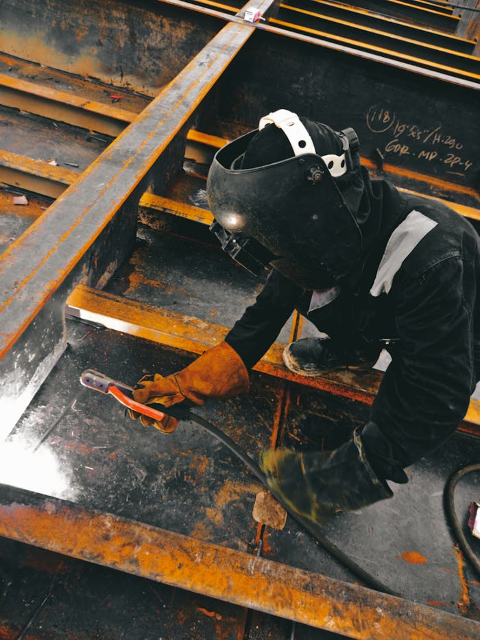 A welder in action wearing protective gear and welding helmet in an industrial setting.